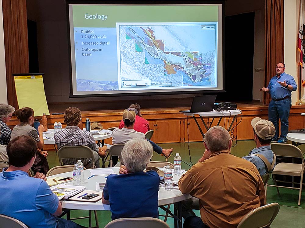 A group of people sitting around several round tables watches a presenter discussing a map of the Cuyama groundwater basin on a large projector screen